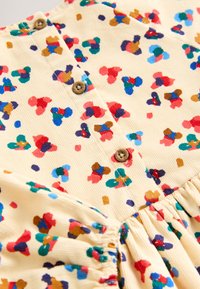Cream fabric dress featuring multicolored abstract spots in red, blue, green, and brown, with wooden buttons and gathered skirt details.