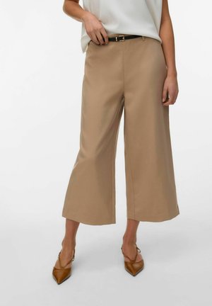 Mujer vistiendo pantalones anchos beige recortados con un cinturón negro, top blanco sin mangas y tacones marrones de punta puntiaguda, de pie contra un fondo liso.