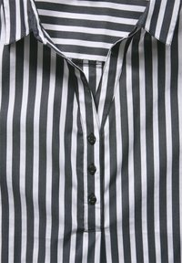 Camicia a righe bianca e nera con colletto, quattro bottoni neri e una patta con dettagli di cucitura visibili.