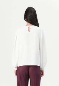 Lightweight white blouse with a keyhole detail at the back, gathered sleeves, and a textured finish. Paired with pleated burgundy pants.