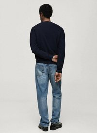 Sweatshirt della marina militare con maniche lunghe, abbinato a jeans blu chiaro. L'individuo è in piedi con le mani dietro la schiena, indossando scarpe nere.