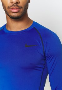 Blaues langärmliges Sportshirt aus glattem, dehnbarem Stoff, mit schwarzem Nähten und einem kleinen schwarzen Nike-Logo auf der oberen linken Brust.