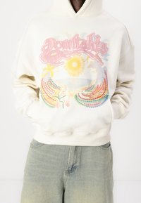 Felpa crema con una grafica frontale che presenta disegni di sole e onde in colori pastello. Polsini e orlo a coste, vestibilità rilassata.