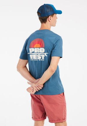 Camiseta de algodón azul con un gráfico retro multicolor en la parte de atrás. Combinada con pantalones cortos coral, modelo con una gorra azul, de pie con las manos entrelazadas.