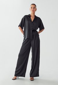 Camicia nera in satin con maniche corte e chiusura a bottoni, abbinata a pantaloni neri in satin a gamba larga. Modella che indossa sandali con tacco.