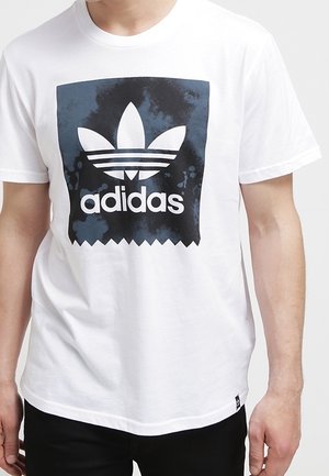 Valkoinen puuvillainen t-paita, jossa suuri musta adidas-logo sinisen liukuvärisen suorakulmaisen taustan päällä, jossa on sahalaitainen reuna.