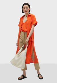 Oranges Hemd mit Knöpfen, passendes Oberteil, beigefarbene, taillierte Hose und schwarze Sandalen. Eine weiße Tasche mit strukturierten Riemen akzentuiert das Outfit.