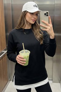 Młoda kobieta w czarnej bluzie i białej czapce trzyma zielony mrożony napój, robi sobie selfie w nowoczesnej windzie z metalowymi ścianami.