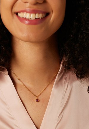 Goldene Halskette mit einem kleinen runden roten Edelsteinanhänger, getragen mit einer blassrosa Bluse. Glatte Textur und filigranes Kettendesign.
