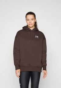 Brun hettegenser med en løs passform, frontlomme og hvit logo aksent. Sammen med svarte leggings med en glatt tekstur.