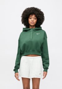 Sweat à capuche court vert avec capuche à cordon, patte de boutonnage et logo Nike blanc. Assorti à un short gris clair texturé.