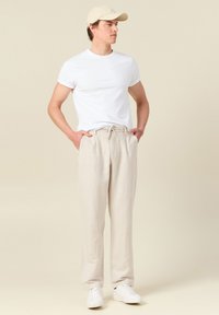 Beiga linnbyxor med dragsko i midjan, kombinerade med en vit bomullströja och vita sneakers, buren av en stående modell.