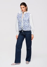Femme portant un chemisier blanc à col et poignets volantés, un gilet bleu à motif floral, un jean foncé et des ballerines transparentes bleues, debout devant un fond uni.