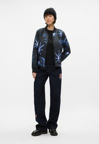 Veste aviateur noire avec des motifs abstraits bleus, associée à un t-shirt noir et un jean en denim sombre ample avec des accents de broderie florale.