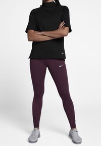 Osoba stojąca z założonymi rękami, ubrana w czarną krótką bluzę z kapturem, bordowe legginsy Nike i szare buty sportowe na białym tle.