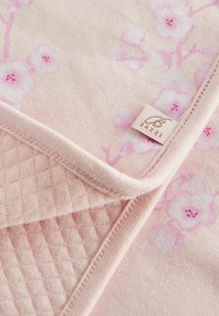 Pătură din bumbac de culoare roz deschis, cu un model floral. Prezintă o textură matlasată pe o parte și un etichete cu logo cusută la margine.