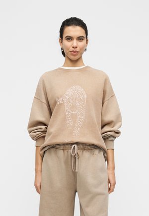 Beżowa bluza z grafiką lamparta, o luźnym kroju i ściągaczami na rękawach. Do tego pasujące beżowe spodnie dresowe z elastycznym pasem.