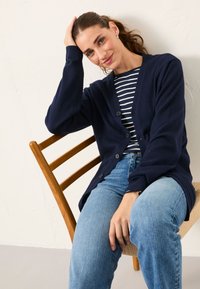 Frau mit braunen Haaren im Pferdeschwanz, die eine marineblaue Strickjacke und ein gestreiftes Hemd trägt, sitzt auf einem Holzstuhl, stützt den Kopf auf die Hand und lächelt.