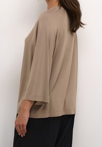 Žena s dlouhými tmavými vlasy, oblečená v volné béžové dlouhé tričko s dlouhým rukávem a černými kalhotami, stojící bokem proti bílé zdi.