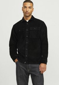 Black corduroy shirt with a collar, two front pockets, and button closure. Textured fabric and slightly loose fit. Worn with dark jeans.
