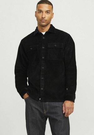 Black corduroy shirt with a collar, two front pockets, and button closure. Textured fabric and slightly loose fit. Worn with dark jeans.