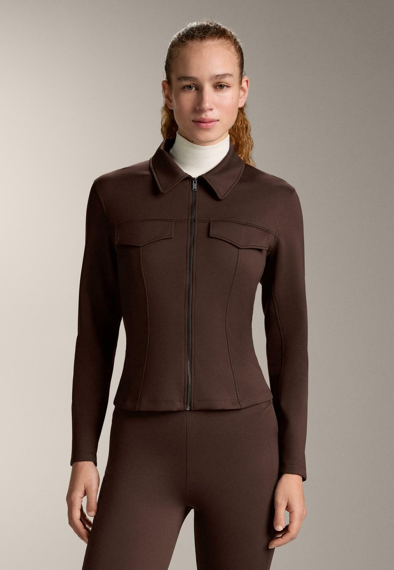 Femme aux cheveux attachés, portant une veste zippée marron foncé cintrée et un pantalon assorti sur un col roulé blanc, debout devant un fond uni.