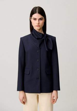Femme aux cheveux raides et foncés portant un blazer bleu marine à fines rayures avec un grand col foulard noué et un pantalon beige sur un fond uni.