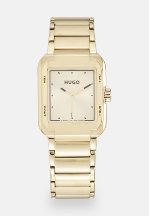 Kuldse tooniga ristkülikuline kell, millel on poleeritud metallist rihm, helesinine nägu, mustad tunni markerid ja brändi nimi "HUGO" selgelt välja toodud.