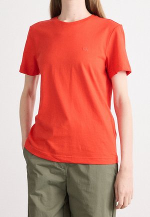 Kobieta ubrana w jednolity, jaskrawy pomarańczowy t-shirt z krótkim rękawem i oliwkowe zielone spodnie, stoi z rękami w kieszeniach na białym tle.