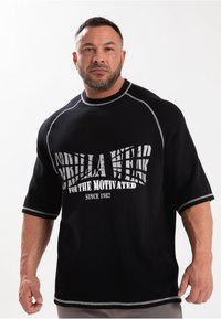 Zwart t-shirt met korte mouwen met witte grafische tekst "Gorilla Wear for the Motivated Since 1982" en contrasterende zilveren stiksels.