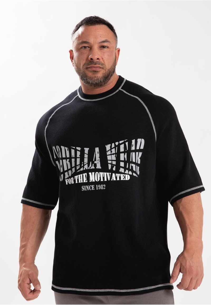 Zwart t-shirt met korte mouwen met witte grafische tekst "Gorilla Wear for the Motivated Since 1982" en contrasterende zilveren stiksels.