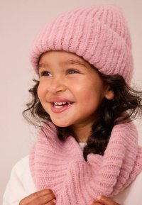 Helle rosa gerippte Strickmütze und passender Schal. Weiche Textur, enganliegende Passform, entworfen, um warm zu halten. Das Modell lächelt, während es die Accessoires trägt.