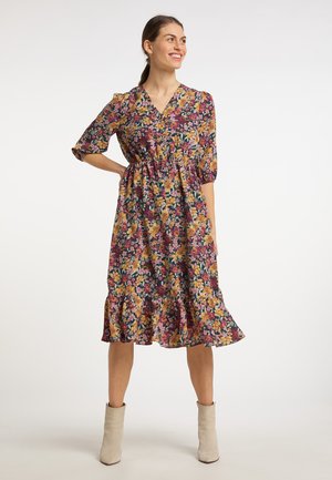 Vrouw staat met hand op heup, draagt een halflange bloemenjurk met pofmouwen en beige enkellaarsjes, glimlacht en kijkt naar rechts.