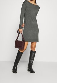 Abito grigio a maglia con maniche lunghe, scollatura a spalla scoperta, abbinato a una borsa bordeaux e stivali neri al ginocchio con tacco.