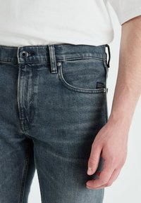 Jeans blu scuro con finitura sbiadita, design a cinque tasche, bottoni metallici e una piccola etichetta sulla tasca anteriore. Il materiale sembra denim.