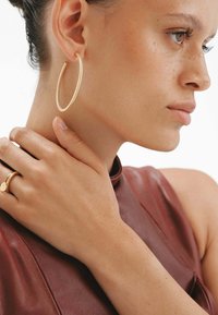 Pendiente de aro de oro con superficie texturizada que presenta un diseño retorcido. El modelo lleva una blusa de cuero marrón y un anillo de oro en su mano.