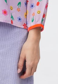 Gonna in tessuto a coste lilla abbinata a una camicetta viola trasparente con motivi di fiori colorati e smile, accentuata da un polsino arancione brillante.