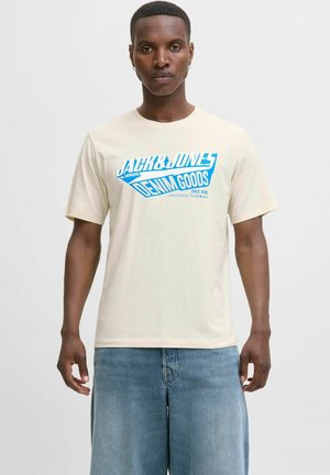 Béžové tričko z bavlny s modrým grafickým designem, na kterém je nápis "JACK & JONES DENIM GOODS" s ostrým stylem. Nosí se s světle modrými šortkami.