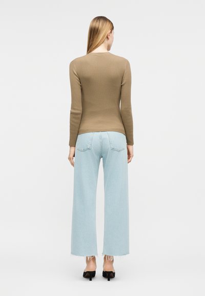 Γυναίκα με πλάτη, φοράει καφέ μακρυμάνικη μπλούζα, ανοιχτό μπλε τζιν παντελόνι με φαρδιά μπατζάκια και ξέφτια στο τελείωμα, και μαύρα σανδάλια με τακούνι.