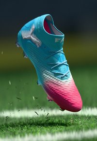 Botte de football bleu vif et rose avec des lacets et des crampons, soulevant de l'herbe sur un terrain de sport.