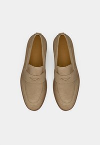 Beige Wildleder-Lederschuhe mit einem Mokassin-Design, das eine genähte Mokassin-Spitze und elastische Seitenpaneele aufweist. Eine braune Gummisohle sorgt für einen Kontrast.