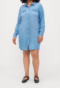 Jasnoniebieska sukienka koszulowa z dżinsu, z długimi rękawami, zapinana na guziki, z dwoma kieszeniami na piersi, długości do kolan, z prostym kołnierzykiem, w zestawieniu z ciemnymi butami z ozdobnymi ćwiekami.