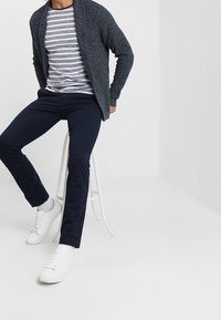 Navy knit cardigan over a gray and white striped T-shirt, paired with dark trousers and white sneakers, seated on a white stool.