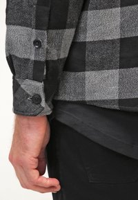 Camisa de franela a cuadros en negro y gris con puños abotonados, que presenta una textura suave y costuras visibles. Debajo, se puede ver una camisa oscura.