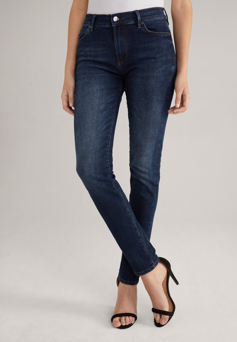 Frau trägt dunkelblaue Skinny-Jeans und schwarze Sandalen mit hohen Absätzen, steht mit gekreuzten Beinen vor einem einfarbigen Hintergrund.