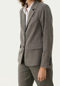 Femme portant un blazer gris texturé à deux boutons et un pantalon assorti, avec une chemise blanche à col en dessous, sur un fond uni.