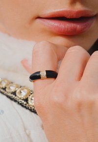 Bague noire au design lisse et incurvé, ornée d'un bandeau en or avec trois petites pierres claires ; portée sur une main de teint clair.