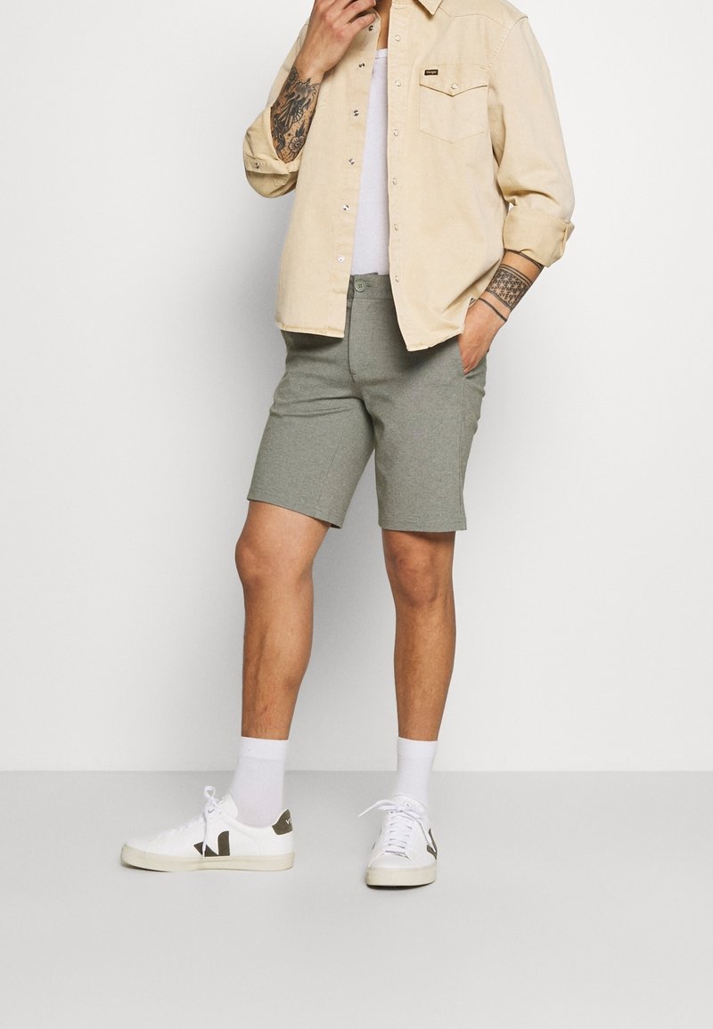 Homme portant une chemise beige à boutons par-dessus un débardeur blanc, un short gris, des chaussettes blanches et des baskets blanches, avec des tatouages visibles sur les deux avant-bras.