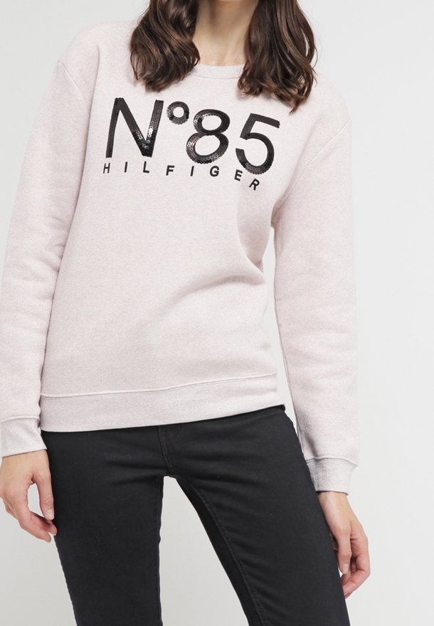 Femme portant un sweat-shirt rose clair avec l'inscription "N°85 HILFIGER" en noir, associée à un pantalon noir, posant devant un fond uni.