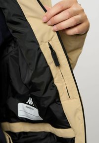 Eine Hand hält eine offene beige Jacke, die ein schwarzes Innenfutter, eine kleine Reißverschlusstasche und ein weißes Etikett mit leerem "Eigentum von"-Text zeigt.